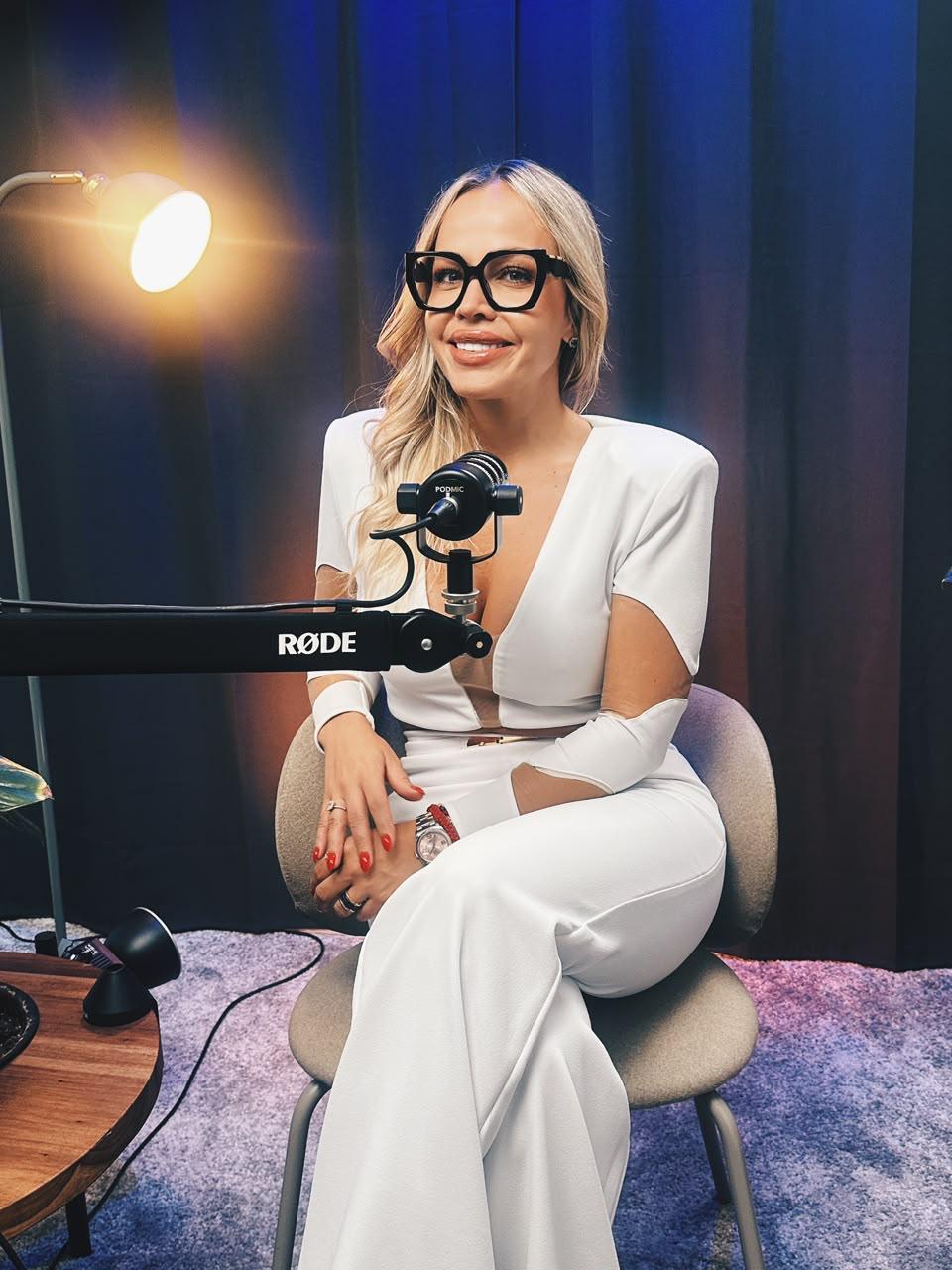 Juli Latorre, sonriente y vestida con un conjunto blanco y lentes de pasta negra, sentada frente a un micrófono profesional durante la grabación de su podcast Al otro lado está la felicidad
